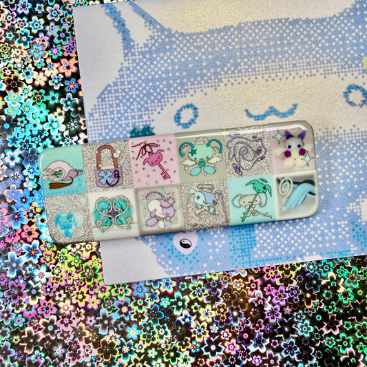 A rectangular hard case with a grid of pastel illustrated panels, placed on top of a blue-and-white patterned sheet over a highly reflective multicolored glitter surface. The case appears to be the product, with decorative fantasy-style artwork including a cat, padlock, key, butterfly, snake, and cloud motifs.