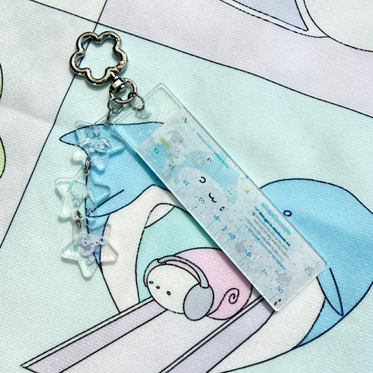 A pastel acrylic keychain featuring a rectangular clear block with printed cute cartoon illustrations and dangling star-shaped charms, shown on a printed fabric background.
