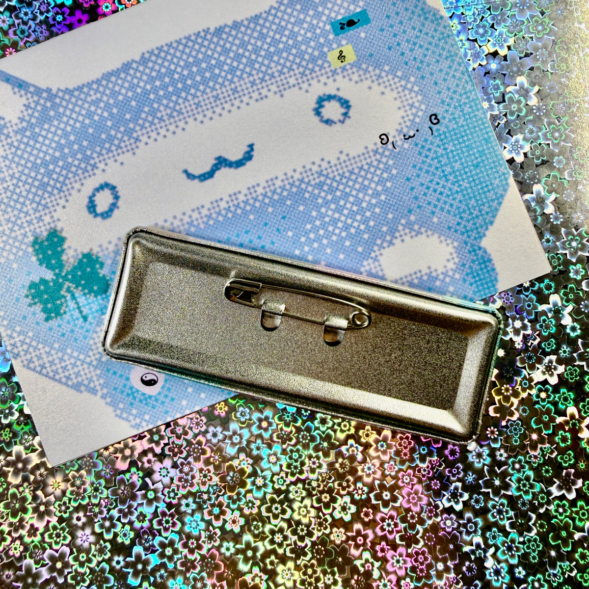 A rectangular metal pin-back badge with a safety pin clasp, shown from the back on top of a blue and white printed card over a holographic glitter surface.