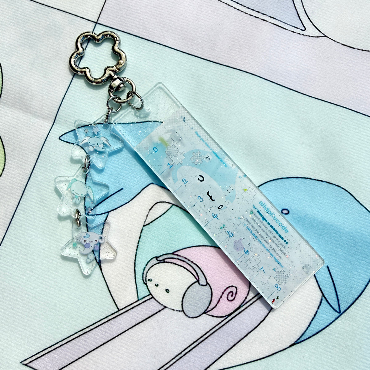 A pastel acrylic keychain featuring a rectangular clear block with printed cute cartoon illustrations and dangling star-shaped charms, shown on a printed fabric background.