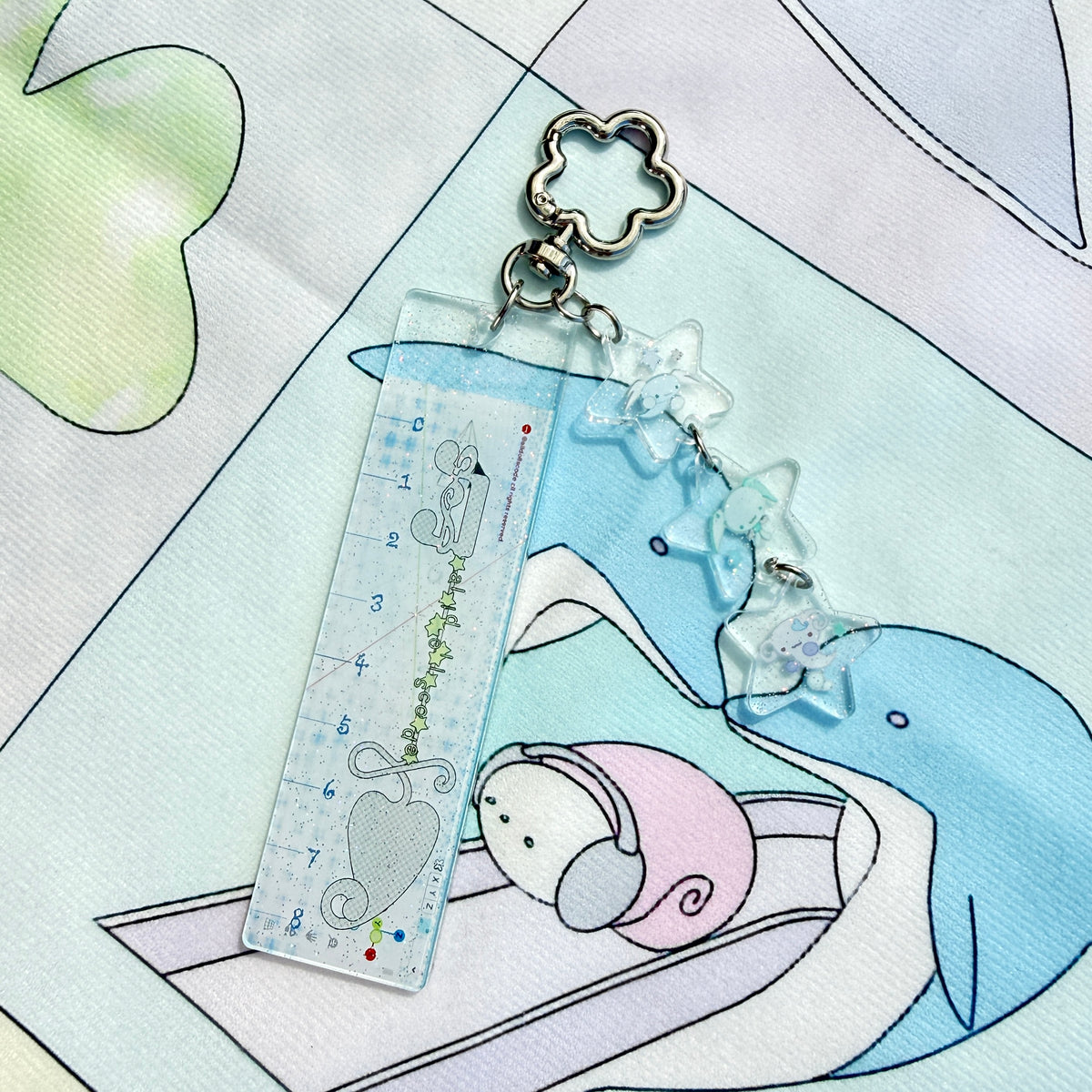 A clear acrylic ruler keychain featuring a cute cartoon mouse and heart design, attached to a silver metal clasp with three clear star charms, resting on a pastel printed fabric.