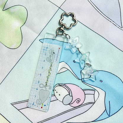 A clear acrylic ruler keychain featuring a cute cartoon mouse and heart design, attached to a silver metal clasp with three clear star charms, resting on a pastel printed fabric.