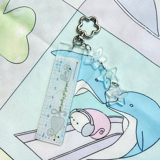 A clear acrylic ruler keychain featuring a cute cartoon mouse and heart design, attached to a silver metal clasp with three clear star charms, resting on a pastel printed fabric.