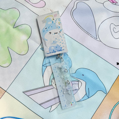 A packaged decorative accessory with a small printed card and a clear plastic tube containing pale blue and white embellishments, placed on a pastel printed fabric background with cartoon animal illustrations.