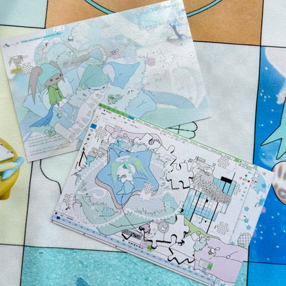 Two printed art cards or stickers with pastel anime-style illustrations, placed on a colorful patchwork mat. One card shows a blue-haired character in a white dress, and the other shows a whimsical illustrated scene with stars and abstract shapes.