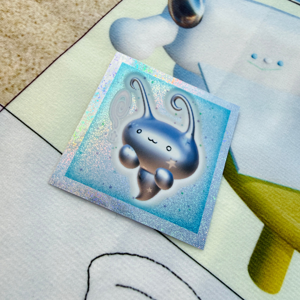 A square holographic sticker featuring a cute metallic blue creature with long curved antennae, placed on a light-colored printed surface.
